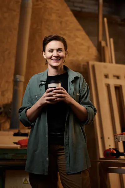 In a vibrant workshop, a talented female carpenter enjoys a moment with her mug, surrounded by wood projects. — Stock Photo
