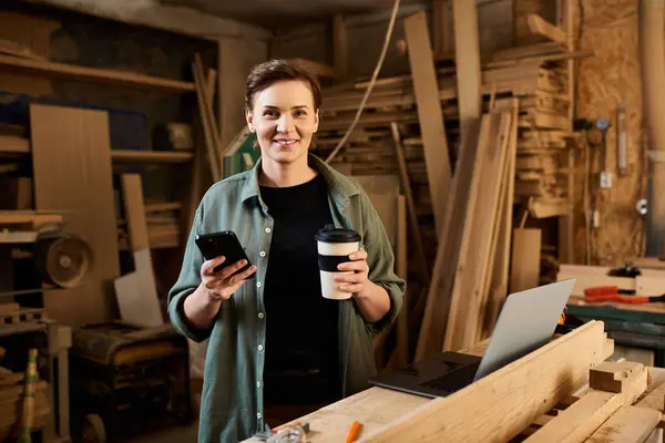 A dedicated carpenter engages in woodworking while balancing coffee and technology in her workshop. — Stock Photo