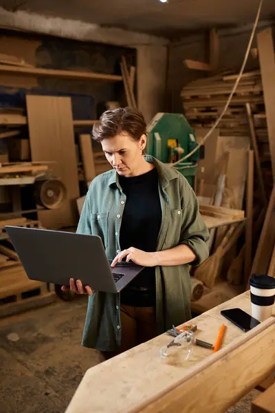 Un experto carpintero revisa los planes de un portátil en un vibrante taller lleno de creaciones y herramientas de madera. - foto de stock