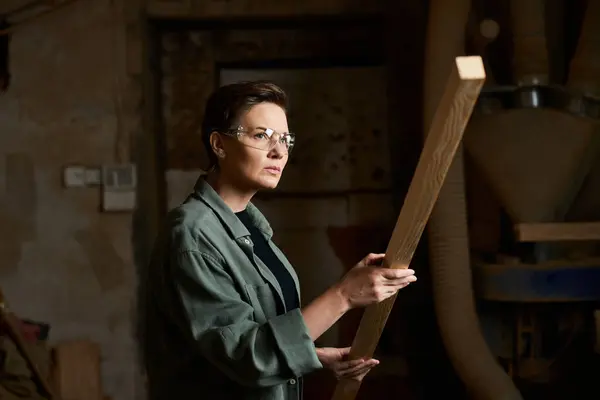 Eine fokussierte Frau hält eine Holzplanke in der Hand, die in ihrer geschäftigen Tischlerei zum Gestalten bereit steht.. — Stockfoto