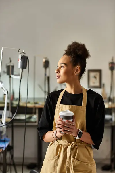 A skilled artisan stands in a workshop, holding a cup of coffee and contemplating her craft. — Stock Photo