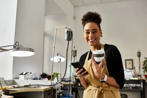 A cheerful woman holds a cup of coffee and a phone while surrounded by jewelry making tools. — Stock Photo
