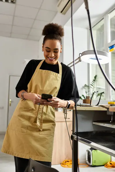 A cheerful woman in an apron happily engages with her phone in a bright, inviting workspace. — Stock Photo