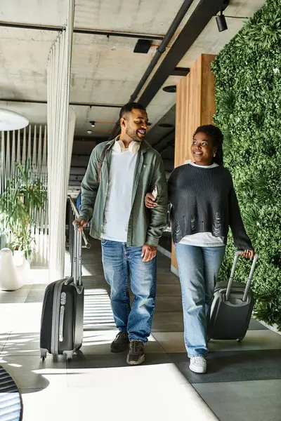 A joyful young African American couple strolls through a vibrant hotel lobby, luggage in hand. — Stock Photo