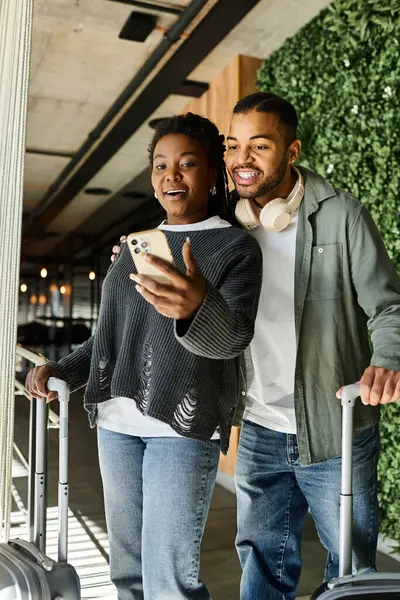 A joyful young couple share a moment in a hotel lobby while checking their travel plans on a phone. — Stock Photo