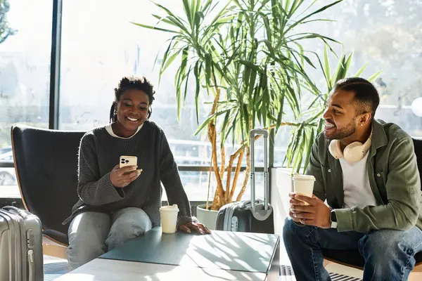 Una alegre pareja joven se relaja en el vestíbulo de un hotel, compartiendo risas mientras mira sus teléfonos. - foto de stock