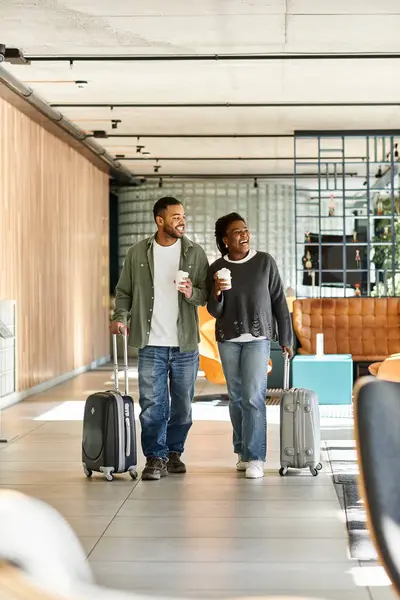 A young couple strolls through a stylish hotel lobby carrying luggage and warm drinks, enjoying their vacation. — Stock Photo