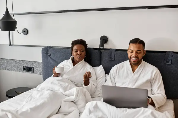 A playful young couple relaxes in robes, using laptop while surrounded by soft bedding and morning light. — Stock Photo