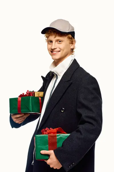 Sporting a trendy winter coat and a cap, a young man beams while holding two beautifully wrapped Christmas gifts. — Stock Photo