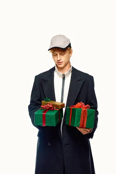 Dressed in trendy winter attire, a young man stands holding two beautifully wrapped Christmas gifts. His fashionable coat and cap reflect the festive spirit. — Stock Photo