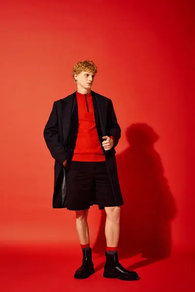 A fashionable man stands confidently in a striking red sweater and stylish black shorts, paired with sleek boots. — Stock Photo