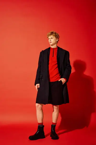 Un hombre con estilo presenta la moda navideña con una acogedora parte superior roja y un elegante abrigo negro. Se apoya con confianza en un fondo rojo vibrante, evocando un espíritu festivo esta temporada de Navidad en 2025. - foto de stock