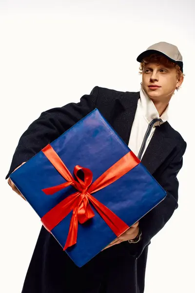 A trendy man showcases a large blue Christmas present wrapped with a bright red ribbon, embodying the festive spirit of 2025. His fashionable winter attire adds to the cheerful atmosphere. — Stock Photo