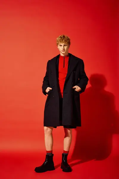 Um homem elegante fica confiante contra um fundo vermelho brilhante, vestindo um pulôver vermelho aconchegante sob um casaco escuro elegante. Seu olhar é completado com botas pretas robustas, irradiando espírito de férias. — Fotografia de Stock