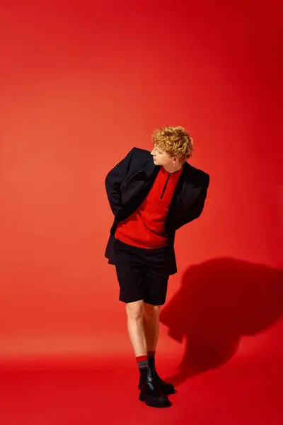 A young man stands against a bold red background, wearing a chic holiday outfit. His ensemble features a striking red sweater paired with tailored black shorts, reflecting modern fashion trends. — Stock Photo