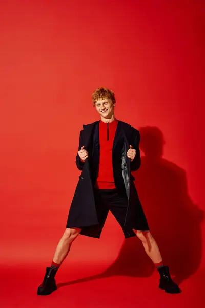 Um homem elegante exibe moda de férias com confiança. Vestido em um vibrante top vermelho e um casaco elegante, ele se coloca contra um impressionante fundo vermelho, incorporando espírito festivo e elegância moderna.. — Fotografia de Stock