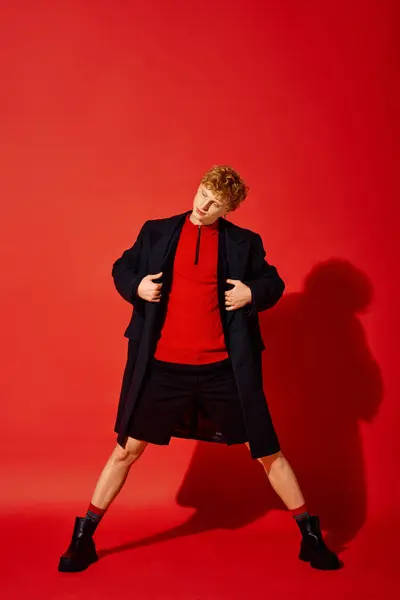 Striking man showcases modern holiday fashion against a vibrant red backdrop. He confidently wears a stylish red top paired with an elegant coat, embodying a fresh take on festive attire. — Stock Photo