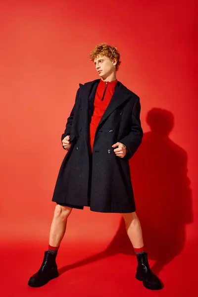 During the Christmas season of 2025, a fashionable man poses confidently in a bold red sweater and striking overcoat. The vivid background enhances his chic look, embodying festive style. — Stock Photo