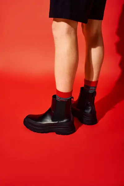 Um homem mostra suas botas pretas na moda emparelhadas com meias vermelhas festivas, de pé contra um fundo vermelho vibrante. Este feriado moda captura a essência do Natal alegria e estilo. — Fotografia de Stock