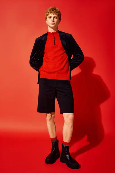 Vestido con un suéter rojo vibrante y pantalones cortos negros a medida, un hombre se levanta con confianza contra un llamativo telón de fondo rojo. — Stock Photo
