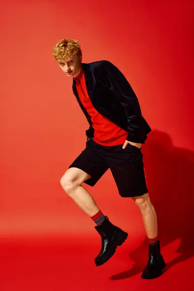 A stylish man showcases the latest holiday fashion against a vibrant red backdrop. He sports a trendy outfit featuring shorts and a sleek jacket, perfect for festive gatherings in winter 2025. — Stock Photo