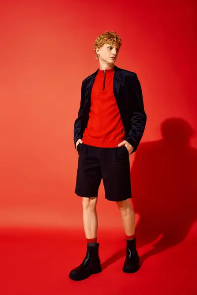 A fashionable man stands confidently against a bright red backdrop, dressed in a striking red top, modern shorts, and trendy boots, showcasing bold style perfect for Christmas 2025 festivities. — Stock Photo