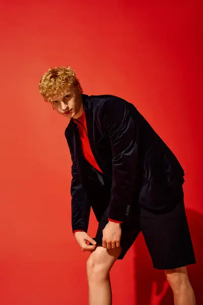 A stylish man showcases unique holiday fashion in a vibrant red setting. He combines a dark blazer with shorts, radiating confidence and creativity for Christmas 2025 celebrations. — Stock Photo