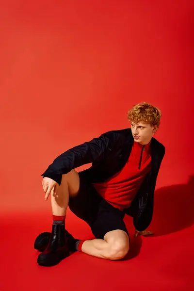 A young man showcases vibrant holiday fashion for Christmas 2025, wearing a bold red top and black shorts. His confident pose against a bright red backdrop exudes seasonal style and flair. — Stock Photo