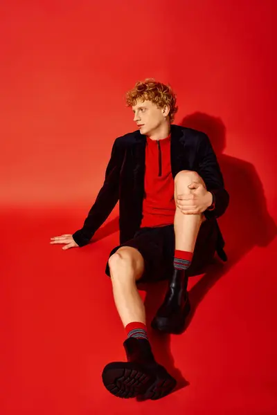 Um homem elegante bate uma pose contra um cenário vermelho vibrante, vestindo um top vermelho na moda, shorts pretos e botas elegantes. — Fotografia de Stock