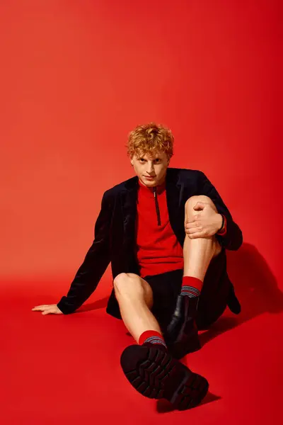 Ein modischer Mann sitzt vor auffallend rotem Hintergrund und strahlt Zuversicht in sein Urlaubsensemble aus. Sein Outfit zeichnet sich durch leuchtende Farben und einzigartige Muster aus, perfekt für saisonale Feiern im Jahr 2025. — Stockfoto
