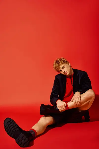 A fashionable man sits casually against a bold red backdrop, displaying modern holiday attire. His ensemble blends comfort and style, perfect for festive gatherings in 2025. — Stock Photo
