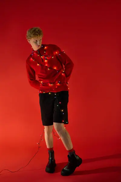 A young man dressed in a cozy red sweater and stylish shorts stands against a vibrant red backdrop. He playfully adorns himself with twinkling Christmas lights, embracing the festive spirit of 2025. — Stock Photo