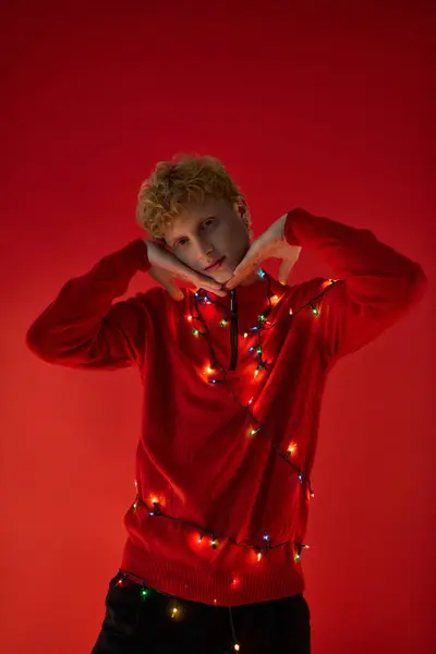 A man poses playfully against a bold red backdrop, draped in colorful Christmas lights. His festive red sweater captures the spirit of the holiday season, reflecting trendy styles for 2025. — Stock Photo
