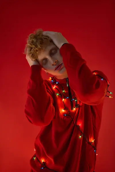 A young man poses stylishly against a bold red backdrop, draped in colorful Christmas lights. — Stock Photo