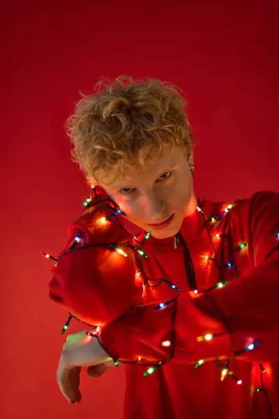 A stylish man showcases vibrant holiday fashion wrapped in colorful Christmas lights against a bright red backdrop. This creative ensemble brings a festive spirit to the holiday season. — Stock Photo
