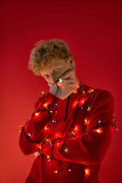 A stylish man is wrapped in colorful Christmas lights, showcasing a playful holiday spirit. The vibrant red background complements his festive outfit, creating a joyful atmosphere for the season. — Stock Photo