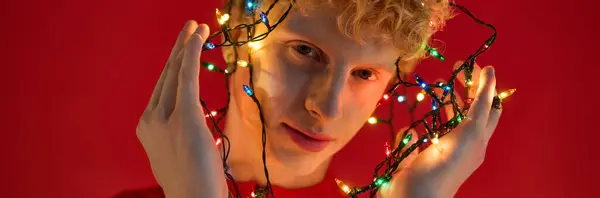 A confident young man flaunts trendy holiday fashion while playfully draping colorful Christmas lights around himself. Set against a vibrant red background, he exudes cheerful holiday spirit. — Stock Photo