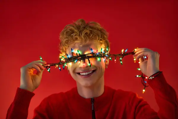 In a vibrant red setting, a young man beams with joy, playfully holding colorful Christmas lights across his eyes. — Stock Photo