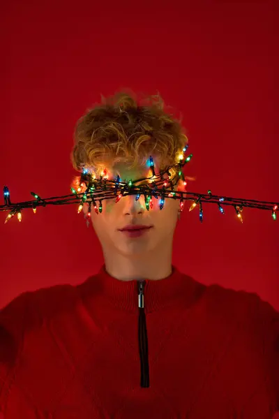 Brightly lit Christmas lights drape across a mans face, embodying the holiday spirit in a striking red setting. His joyful expression showcases his stylish seasonal outfit, perfect for celebrations. — Stock Photo