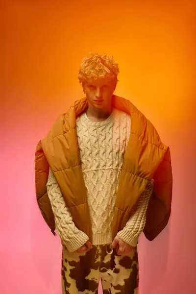 A stylish young man confidently sports a chunky sweater and bold puffer jacket, against a vibrant orange and pink backdrop, capturing the holiday spirit for Christmas 2025. — Stock Photo