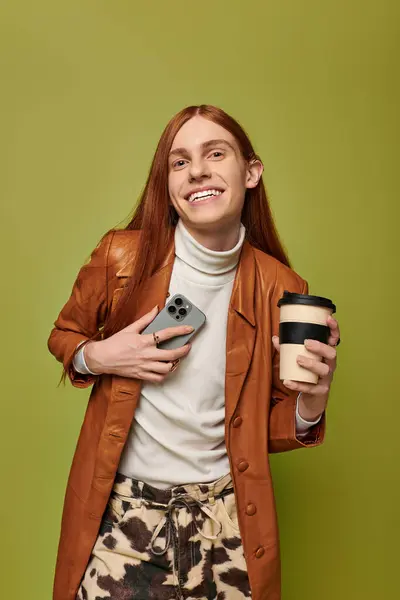 A young man with long red hair beams while holding a coffee cup and phone, radiating happiness. — Stock Photo