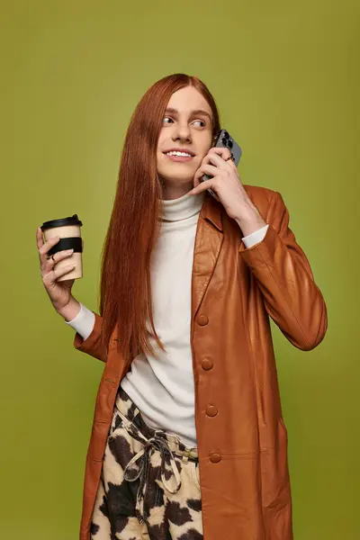 A young man with long red hair smiles while holding a coffee and chatting on the phone. — Stock Photo