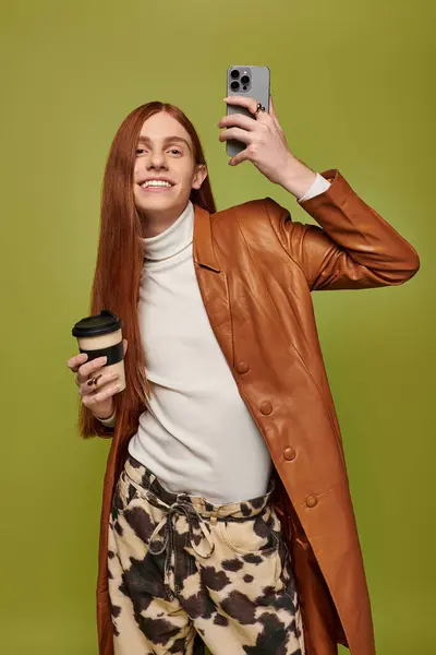 Emotional young man holds coffee and takes a cheerful selfie against a green backdrop. — Stock Photo