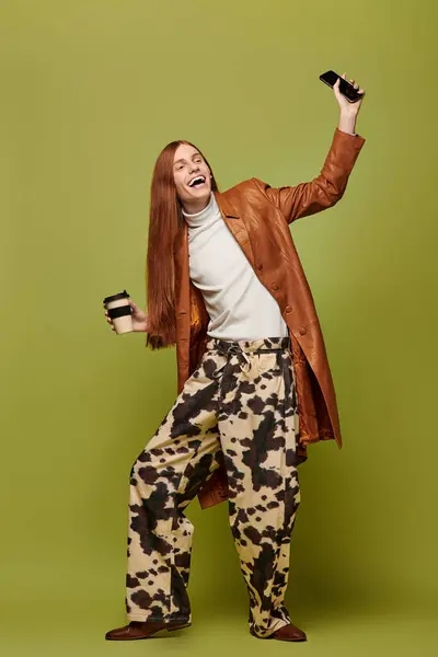 Young man with long red hair wears trendy outfit while holding coffee and smartphone, smiling. — Stock Photo