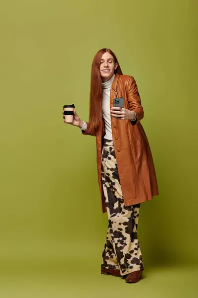 A young man with long red hair expresses joy while holding a drink and a phone. — Stock Photo