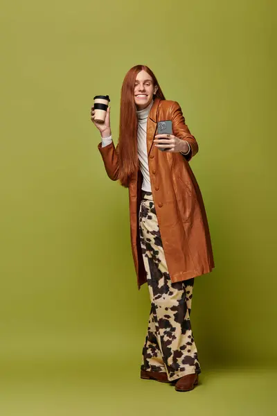 Joyful young man with long red hair holds a coffee cup and phone, radiating confidence and style. — Stock Photo