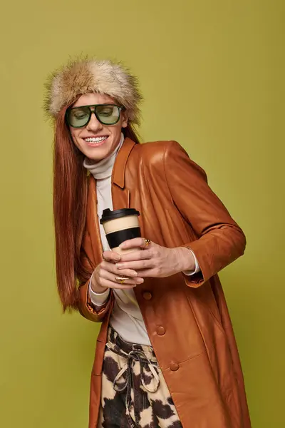 Emotional young man with long red hair smiles brightly while holding a warm drink in cozy attire. — Stock Photo