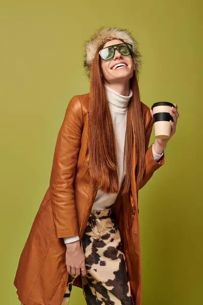 Joven con el pelo rojo largo sonríe alegremente sosteniendo una bebida caliente contra un colorido telón de fondo. - foto de stock