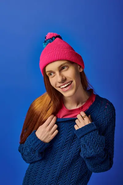 Young man with long red hair smiles in a colorful beanie against a bright blue background. — Stock Photo
