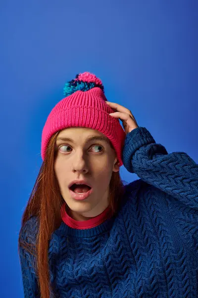 Young man with long red hair looks surprised while wearing a colorful beanie in a lively setting. — Stock Photo